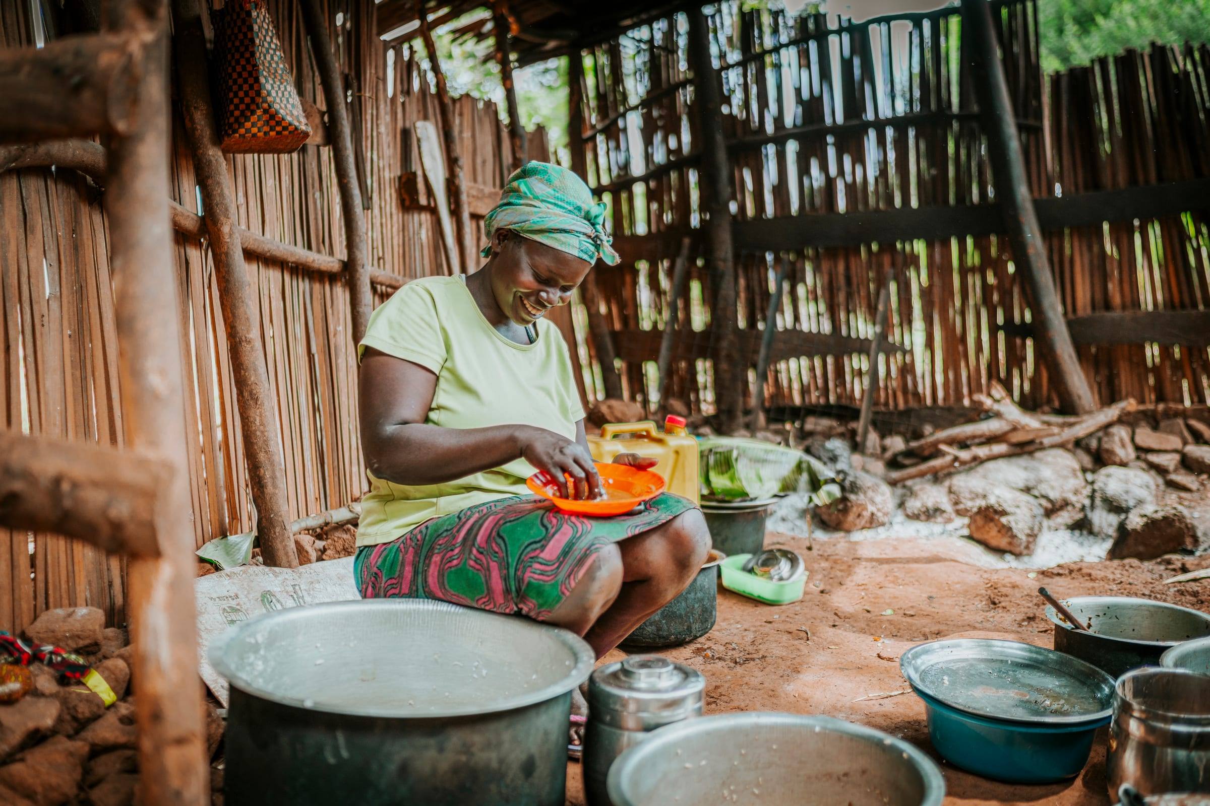 A woman in a green headwrap smiles as she prepares food in an outdoor kitchen
