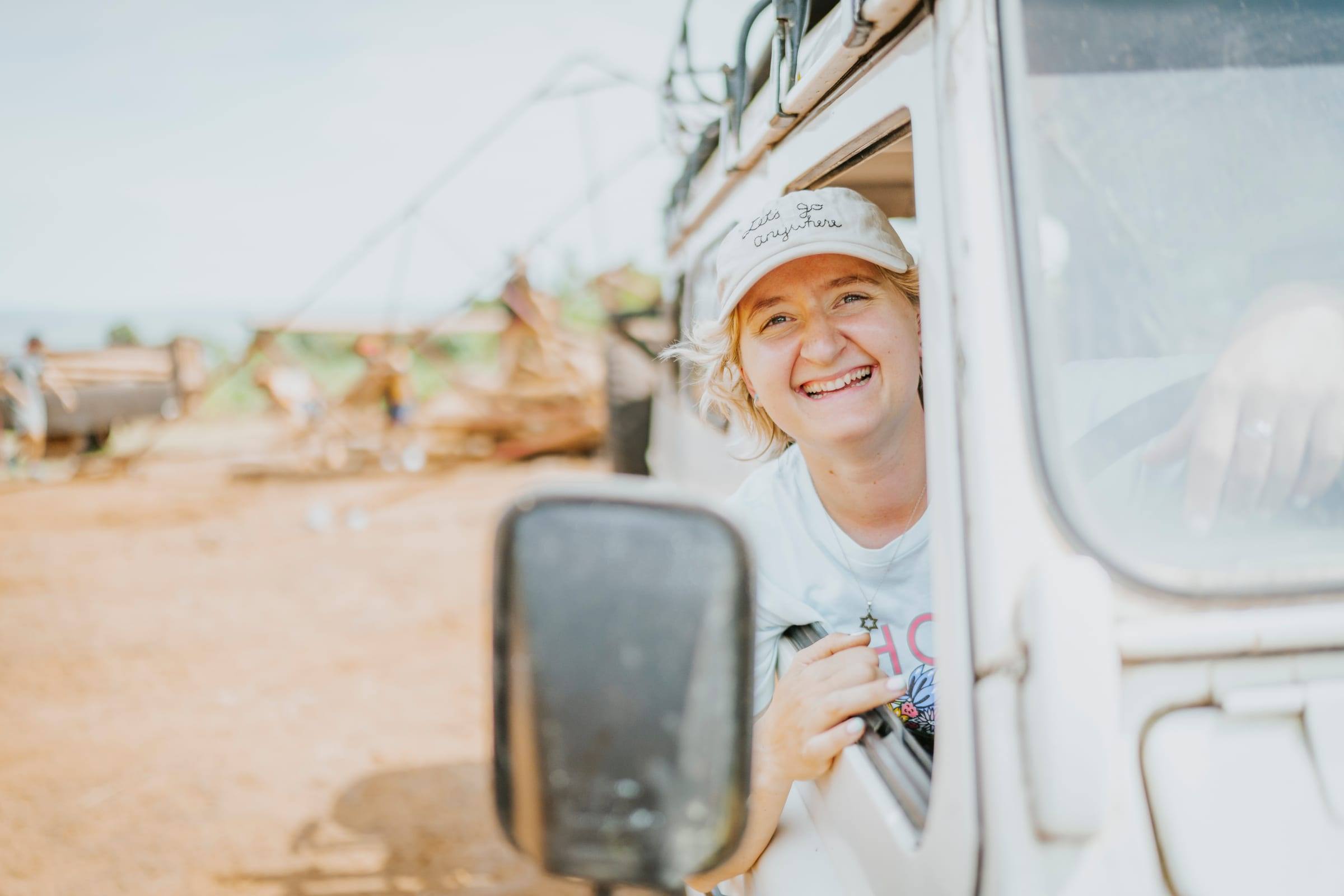 A smiling traveler in a cap leans out of a vehicle window
