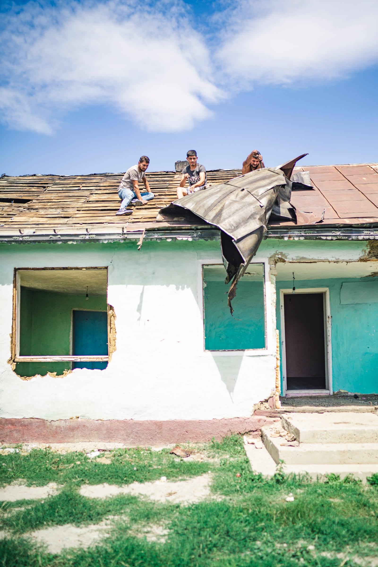 Workers on a roof strip the old roofing from a white house with teal windows