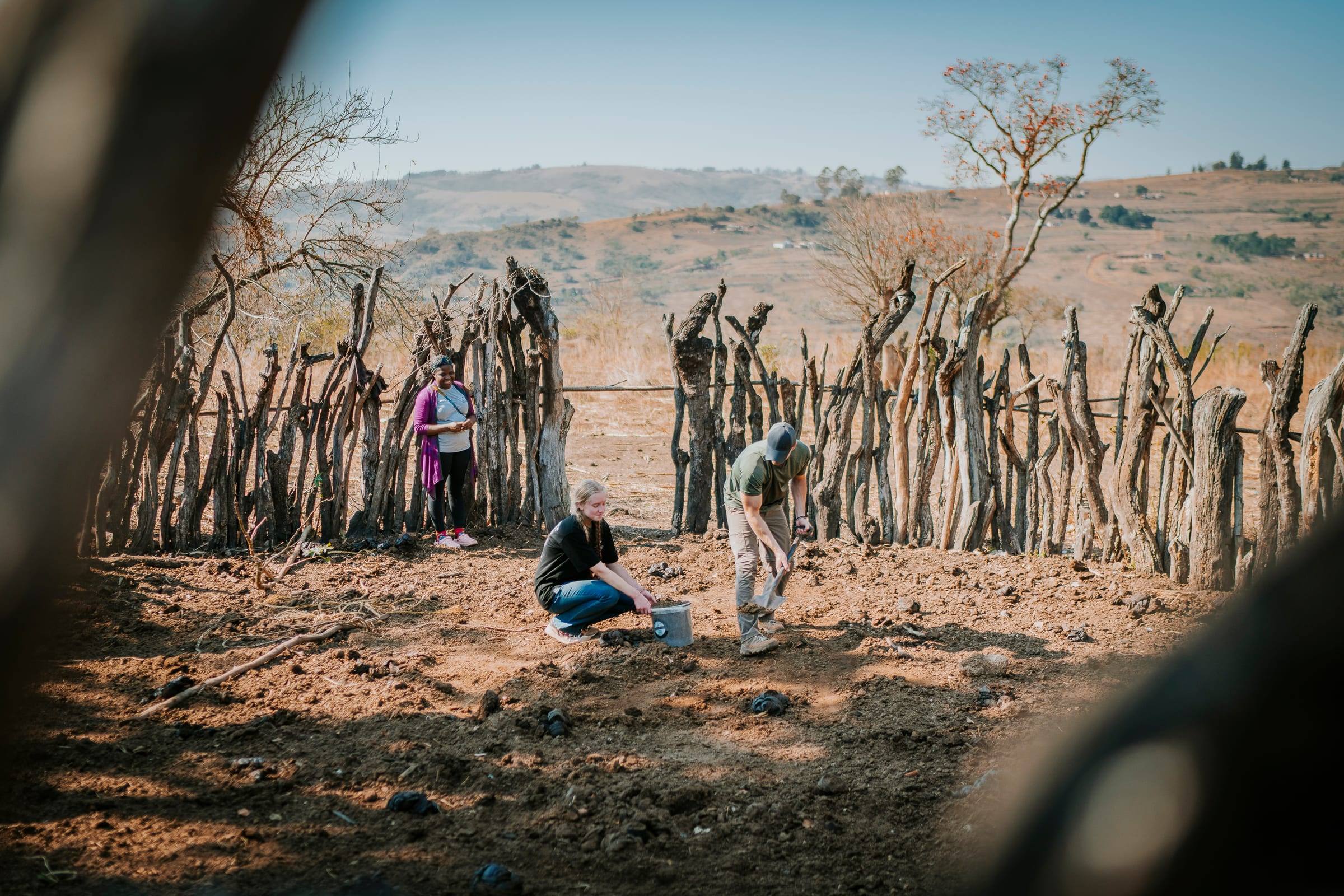 Team members dig inside a fence of wooden stakes with dry hills beyond