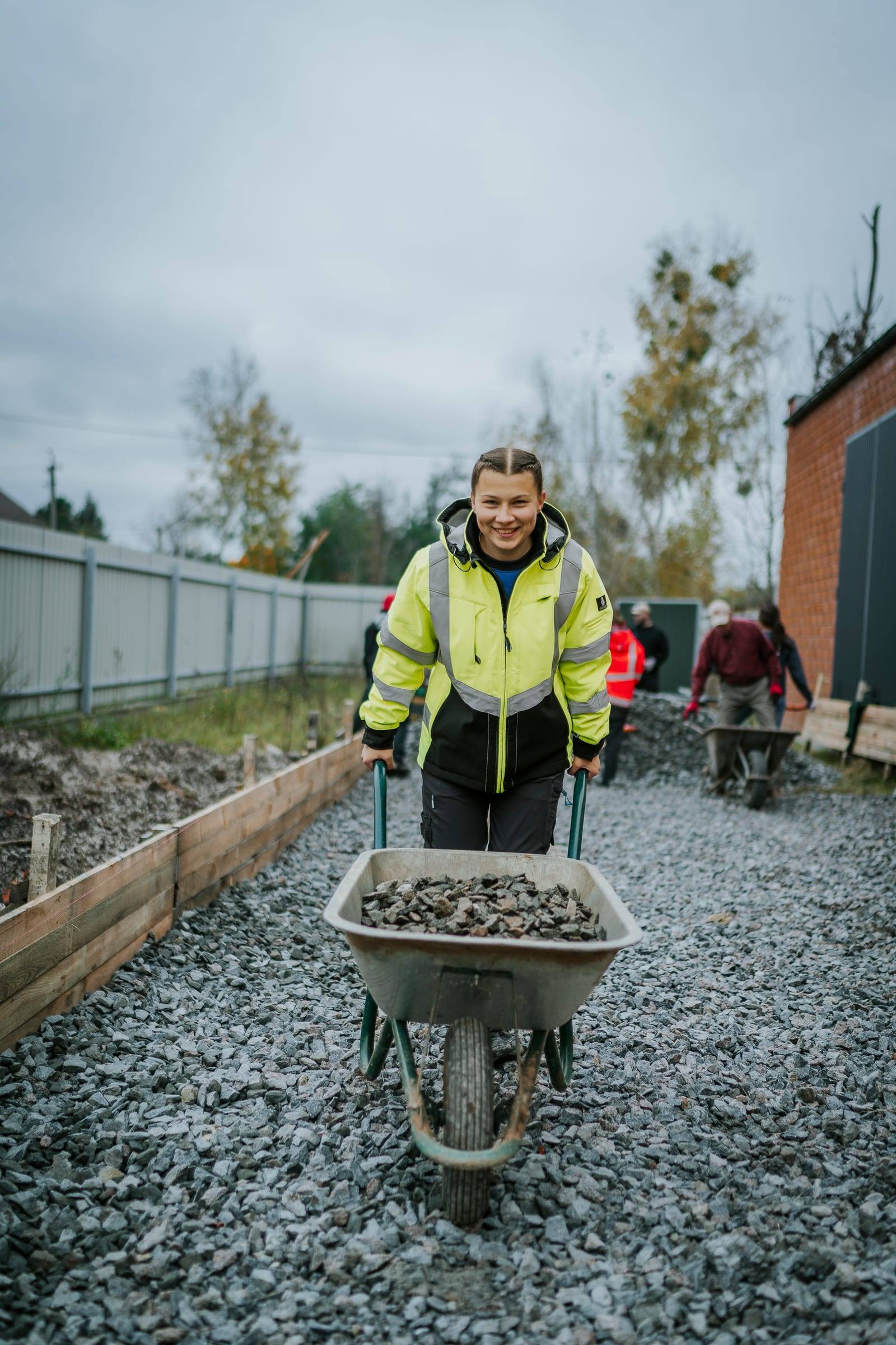 A smiling volunteer in a bright jacket pushes a wheelbarrow of gravel