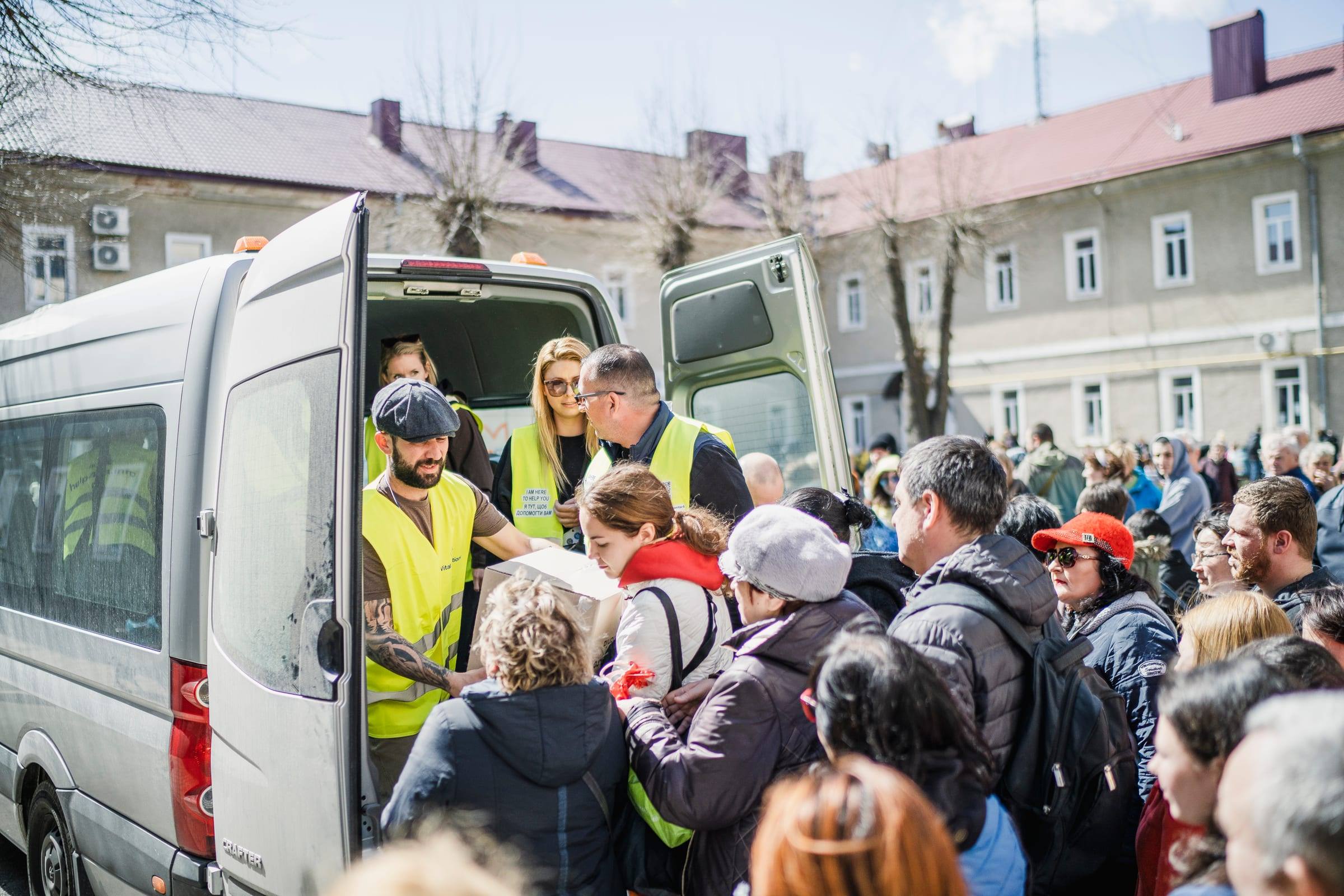 A crowd of families gathers at a van as volunteers hand out food
