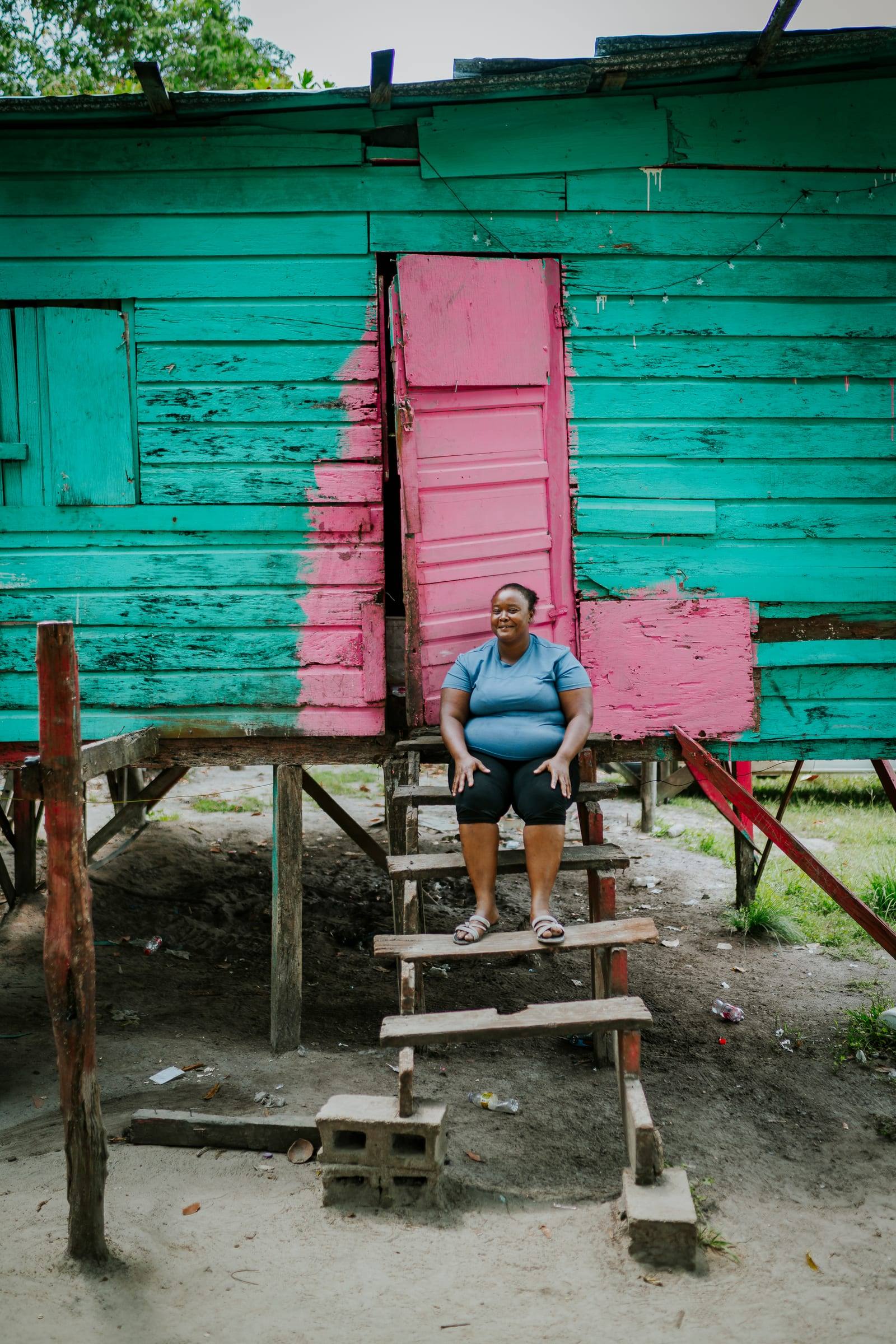 A woman sits on the steps of her teal stilt home with a pink door