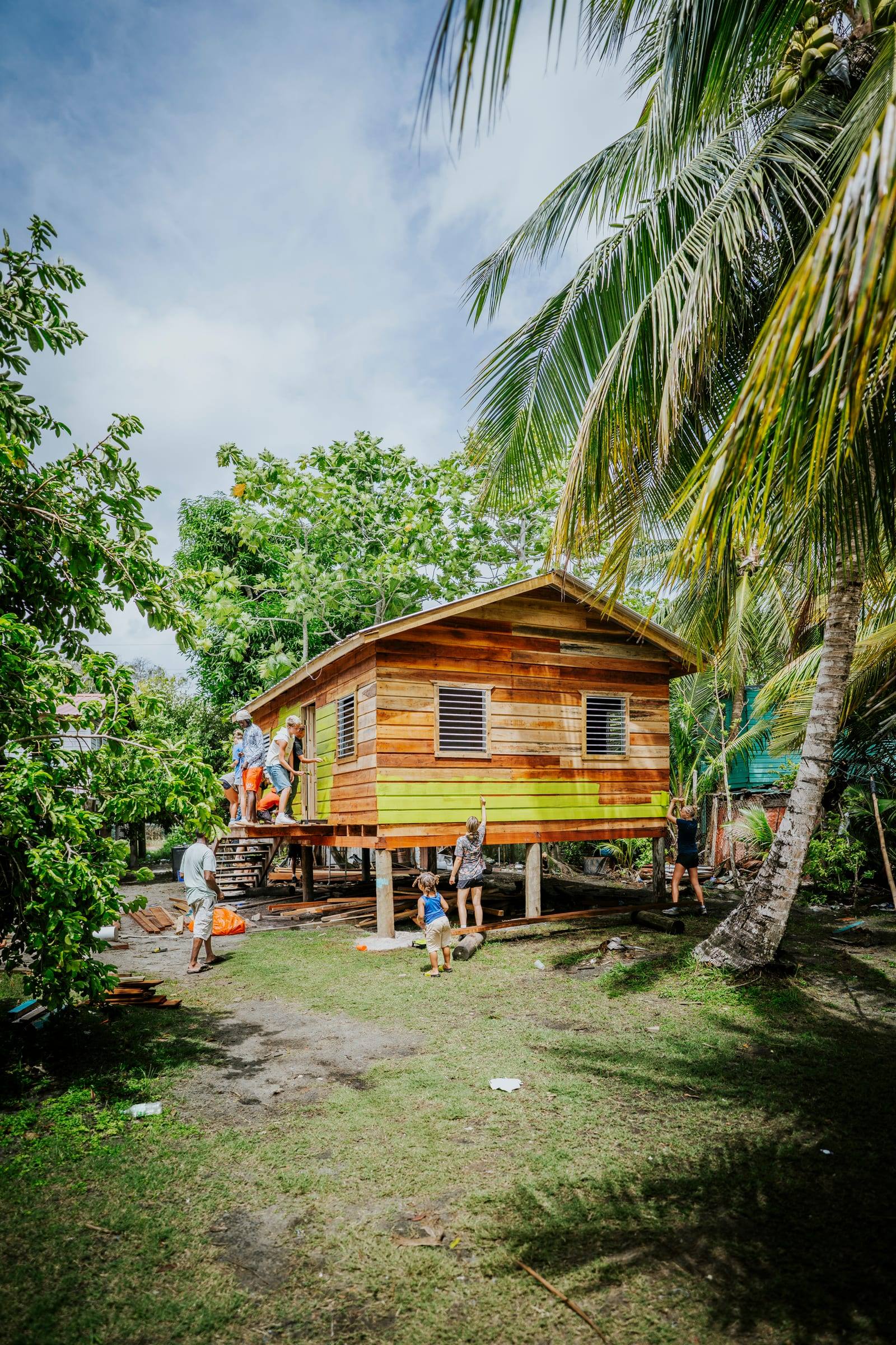 A finished wooden home stands on stilts under palm trees, children playing below