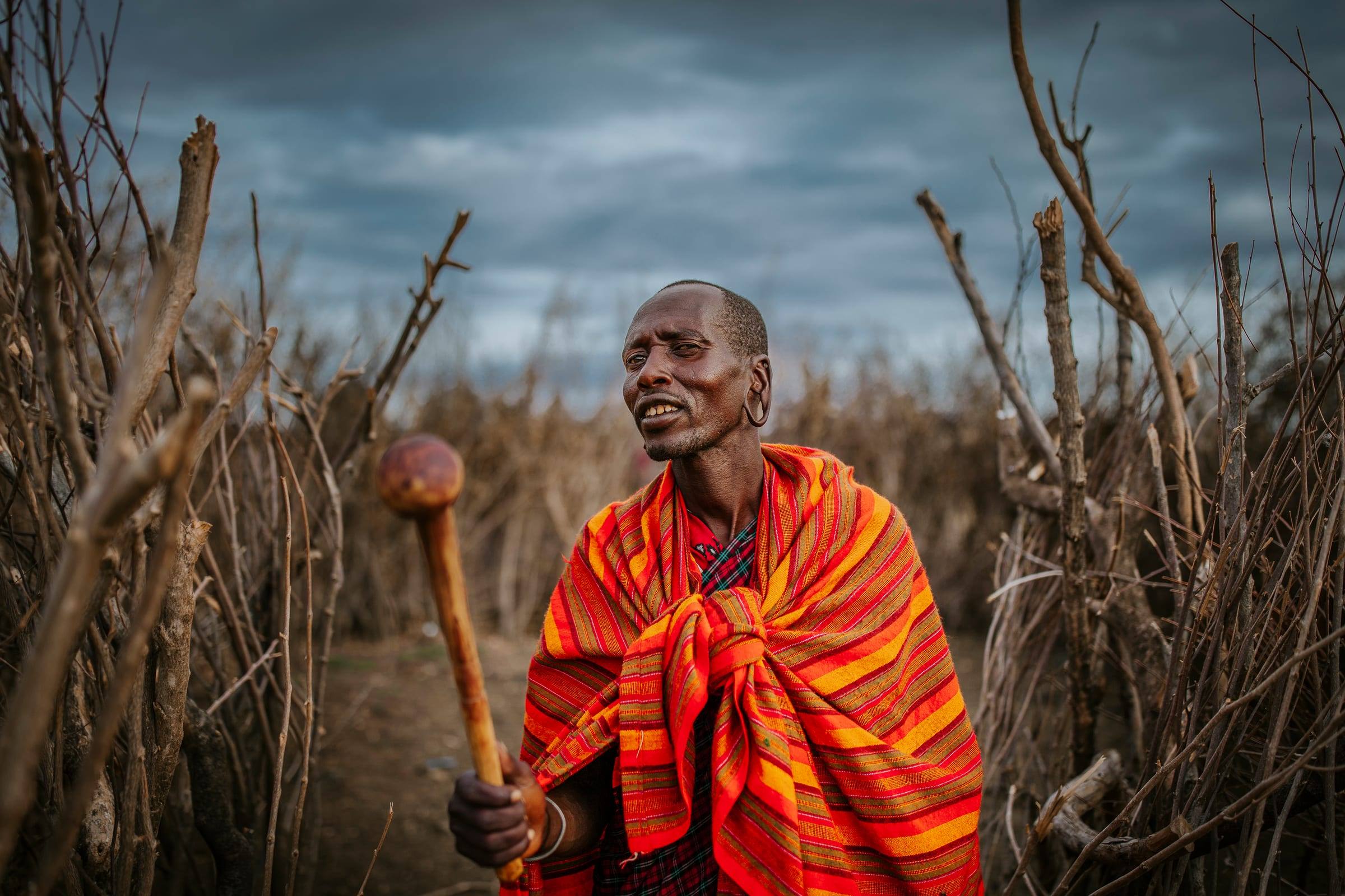 A man in a bright red wrap stands in a field of dry maize stalks under a stormy sky