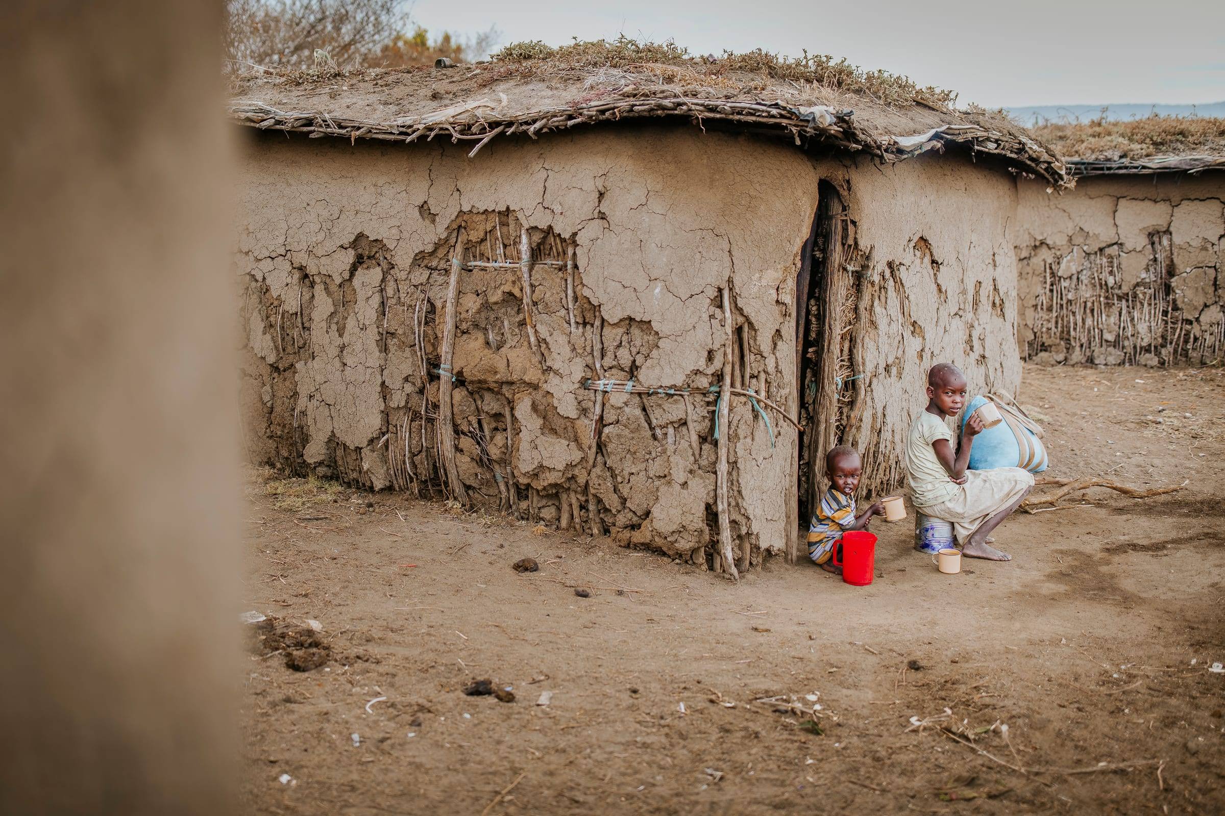 Two children sit at the doorway of a mud walled homestead