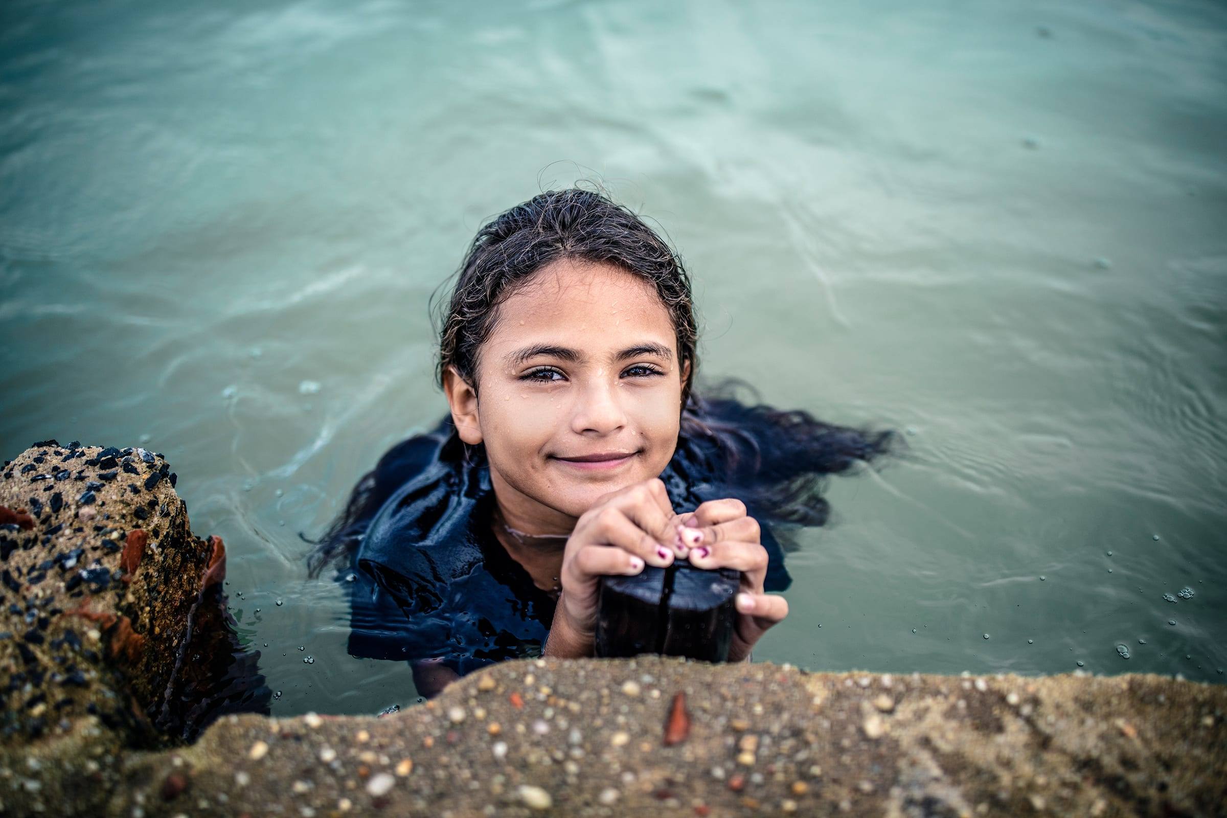 A girl smiles up from the water beside a rock ledge