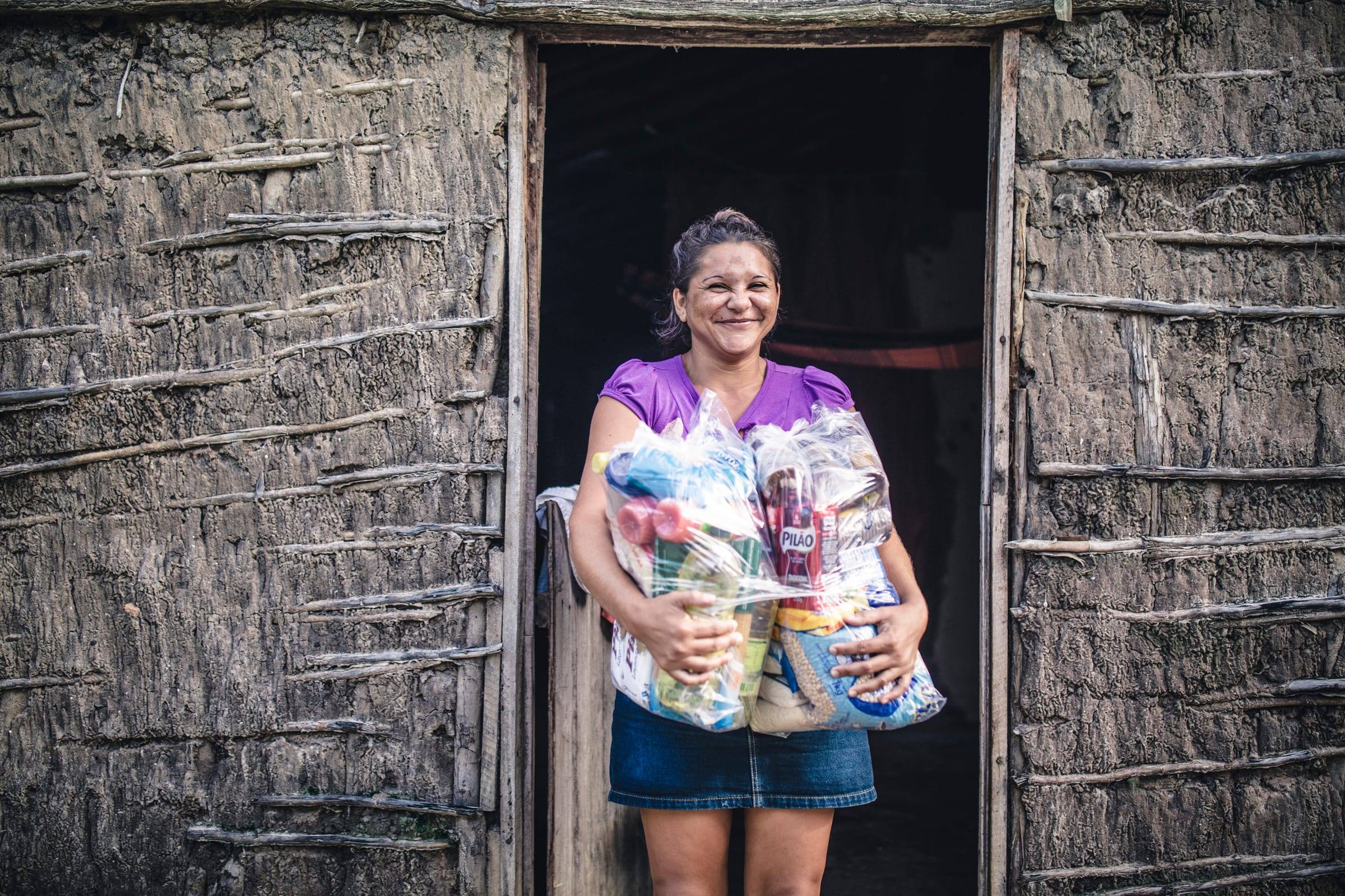 A woman smiles in the doorway of her home holding bags of food staples